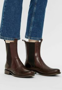 Bottines en cuir marron avec des panneaux élastiques sur les côtés, texture lisse, bout arrondi et petit talon empilé. Un pantalon en denim est visible au-dessus.