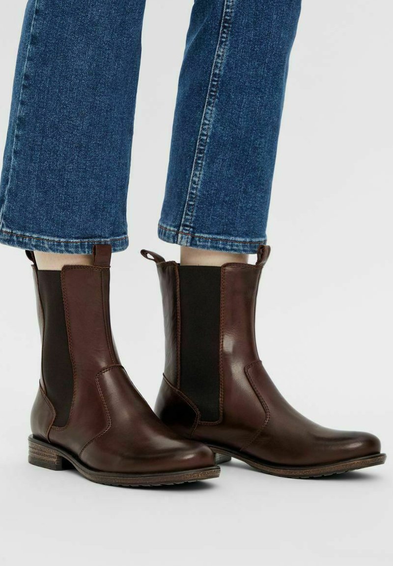 Bottines en cuir marron avec des panneaux élastiques sur les côtés, texture lisse, bout arrondi et petit talon empilé. Un pantalon en denim est visible au-dessus.