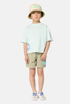 Licht muntgroen T-shirt met korte mouwen en een afbeelding aan de zoom, gecombineerd met olijfgroene shorts met een patch. Crèmekleurige schoenen met blauwe veters. Emmerhoed.