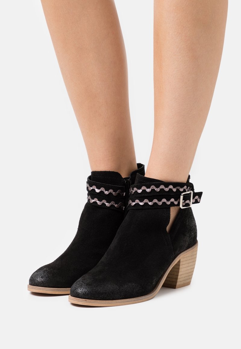 Bottines en daim noir avec talons en bois empilés, bride ondulée décorative avec boucle en argent autour des chevilles, portées sur des jambes nues.