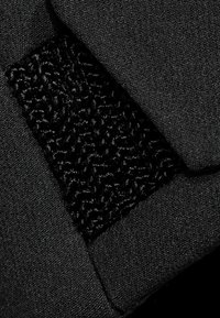 Tissu noir avec un patch texturé présentant une surface pelucheuse et surélevée. Le matériau est lisse et contraste avec la zone environnante.