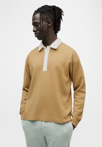 Καφέ πουλόβερ με λευκό γιακά και πατιλέτα, με μακριά μανίκια και άνετη εφαρμογή, συνδυασμένο με ανοιχτοπράσινο παντελόνι από κοτλέ.