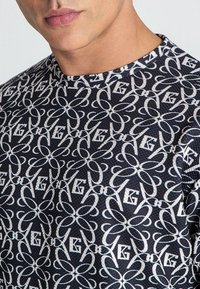 Čierno-biely krátky rukávový sveter s vzorom, ktorý zobrazuje prepletené kvetinové a písmenové motívy v textúrovanej tkanine. Okrúhly výstrih je viditeľný.