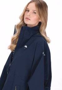 Tengerészkék vízálló dzseki magas gallérral, cipzáras záródással és állítható húzózsinórral. Kis esernyő logóval a mellrészen és rugalmas betétekkel.