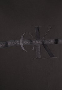 Czarny bawełniany t-shirt z wypukłym logo "CK" i satynową wstążką z napisem "CALVIN KLEIN JEANS" w czerni, na gładkiej, matowej powierzchni.