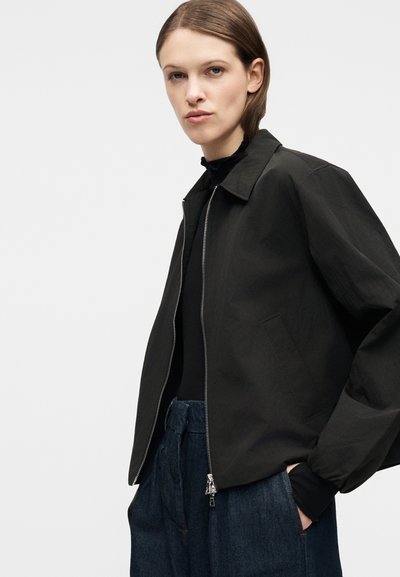 Νεαρό άτομο που φοράει μαύρο μπουφάν με φερμουάρ πάνω από μαύρο τοπ και ψηλόμεσα σκούρα μπλε τζιν, με τα χέρια στις τσέπες, μπροστά σε απλό ανοιχτό φόντο.