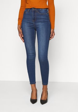 Osoba ubrana w niebieskie obcisłe dżinsy, pomarańczowy sweter z prążkowanego materiału i czarne szpiczaste szpilki, stojąca na białej podłodze.