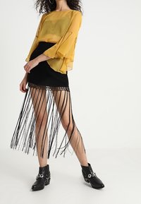 Blouse jaune transparente avec des manches évasées, associée à une jupe noire à franges. Des bottines noires avec des accents métalliques complètent le look.