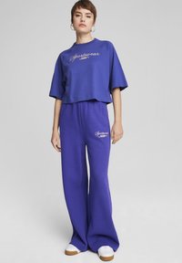 Violetinio medvilninio trumpinio marškinėlių su "Sportswear" užrašu derinys su plačiomis violetinėmis treningų kelnėmis ir baltais sportiniais bateliais. Paprastas, laisvas dizainas.