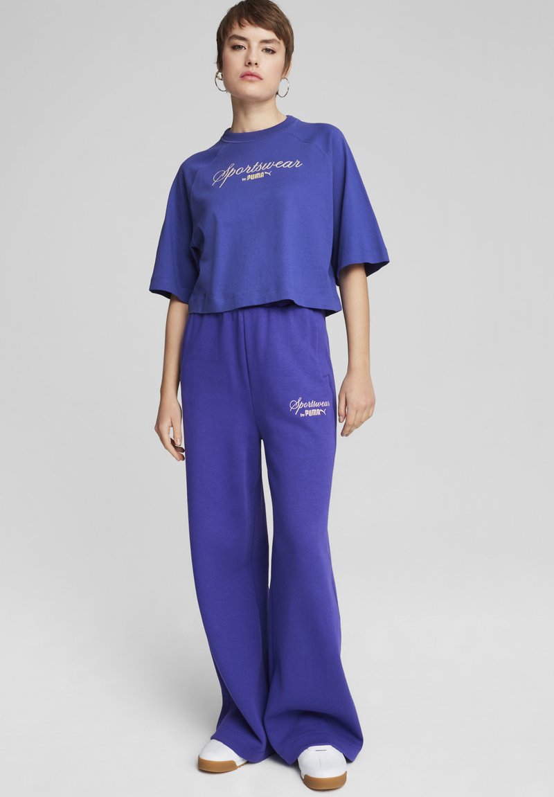 Violetinio medvilninio trumpinio marškinėlių su "Sportswear" užrašu derinys su plačiomis violetinėmis treningų kelnėmis ir baltais sportiniais bateliais. Paprastas, laisvas dizainas.