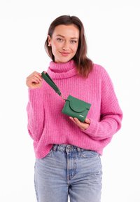 Green leather wallet with a round flap and wrist strap, held by a person in a pink knitted sweater. Features three decorative dots.