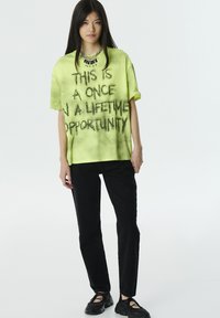 Camiseta oversize de color amarillo neón con texto en negro pintado con spray que dice "ESTA ES UNA OPORTUNIDAD ÚNICA EN LA VIDA", combinada con jeans negros y calzado.