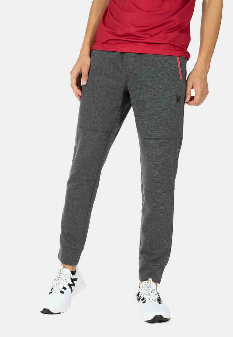 Grå atletiske joggers med en tekstureret finish, der har et sort logo, sidelomme med lynlås og tætsiddende manchetter, sammen med hvide sneakers.