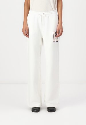 Personne portant un pantalon de jogging blanc à jambes larges avec taille à cordon et un grand patch "H" sur la cuisse gauche, associé à des chaussures noires.