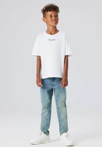 Bela bombažna majica s tekstom "Studio edition", v kombinaciji s svetloplavimi kavbojkami in belimi športnimi copati. Mehak material in sproščen kroj.