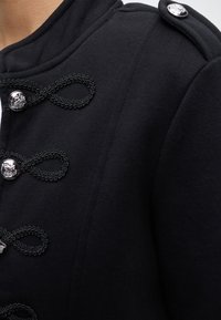 Gros plan d'une veste noire avec des boucles tressées décoratives noires et des boutons argentés à l'avant et une épaulette à l'épaule.