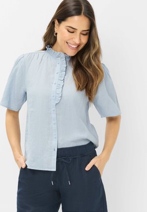 Frau lächelt, trägt eine hellblaue, kurzärmelige Bluse mit Rüschenkragen und dunkelblaue Hose mit Kordelzug, Hände in den Taschen.