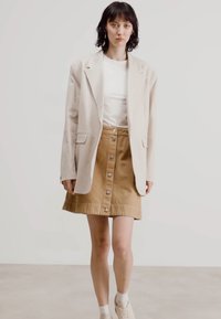 Blazer in lino beige sopra una t-shirt bianca, abbinato a una gonna button-up color beige. La modella è in piedi di fronte a uno sfondo neutro, indossando sneakers bianche.