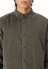 Chemise en velours côtelé vert foncé avec un col rabattu et des boutons noirs contrastants, présentant une texture rayée verticale et des manches longues.