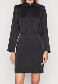 Svart långärmad skjortklänning i ett mjukt tyg. Den har en knappstängning framtill och en figurnära silhuett som betonar en elegant design.