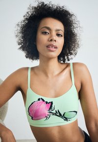 Mintgrön bralette med ett stort rosa blommönster med svarta konturer. Den har tunna axelband och en mjuk, elastisk struktur.