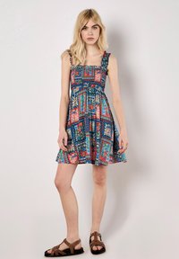 Vestido con estampado floral en azul con detalles en rojo y verde. Presenta tirantes con volantes, un corpiño ajustado y una falda en línea A. Combinado con sandalias marrones.