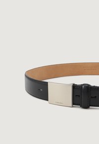 Curea din piele neagră cu o finisare netedă, dotată cu o cataramă metalică dreptunghiulară argintie gravată cu "Calvin Klein." Interiorul este din piele bej.