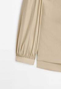 Blouse beige à manches longues avec des poignets froncés et un ourlet droit sur fond blanc.