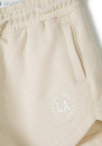 Bermudas de algodón beige con cinturilla elástica, bolsillo lateral y logo impreso "LA" en diseño circular. Textura suave.