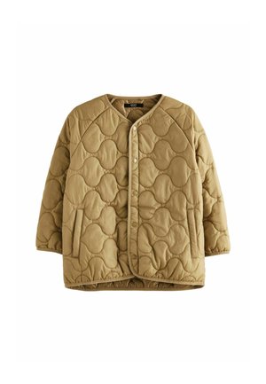 Personne portant une veste matelassée beige avec un motif de couture ondulée, les mains dans les poches, debout contre un mur texturé vert clair.