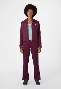 Maroon track jacket with blue side stripes and matching pants, featuring a zip front and logo. Worn over a light blue top and sneakers.