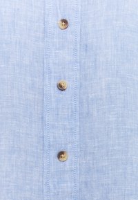 Lichtblauwe linnen overhemd met een voorpaneel voorzien van vier ronde, lichtbruine knopen van kunststof, met een subtiele textuur in de stof.