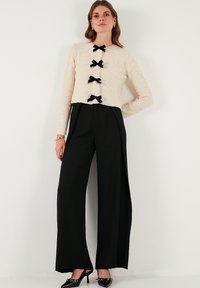 Cardigan in maglia beige con tre accenti neri a forma di fiocco, abbinato a pantaloni neri larghi e scarpe nere a punta. Texture lisce e design aderente.