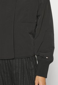 Black cropped jacket made of lightweight fabric, featuring a smooth texture, ribbed cuffs, and a side pocket.