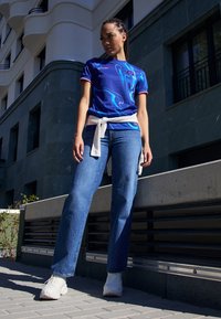 Blå Nike-tröja med abstrakt vitt och blått mönster, röda accenter, i kombination med ljusblå raka jeans och vita sneakers, knuten skjorta.