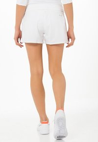 Pantalones cortos deportivos blancos hechos de tela ligera, con un diseño ajustado, una marca sutil y combinados con zapatillas blancas con detalles naranjas.