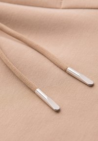 Gros plan sur un tissu beige avec deux cordons beiges se terminant par des embouts métalliques argentés brillants.