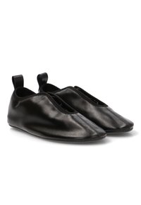 Zapatos de cuero negro tipo mocasín con punta afilada, apertura elástica y pequeñas pestañas en el talón para ponerlos fácilmente. Textura suave y diseño minimalista.