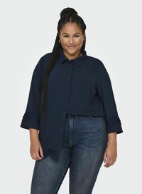 Camicia button-up blu navy con colletto e maniche a tre quarti, caratterizzata da rifiniture in pizzo. Abbinata a jeans in denim scuro.