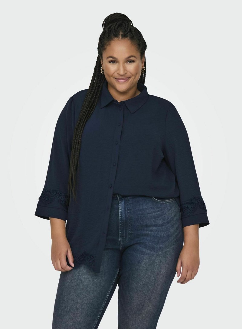 Camicia button-up blu navy con colletto e maniche a tre quarti, caratterizzata da rifiniture in pizzo. Abbinata a jeans in denim scuro.