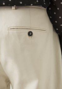 Beiga byxor i texturerat tyg med en bakficka som stängs med en svart knapp. Diskreta sömmar som detaljer längs fickan.