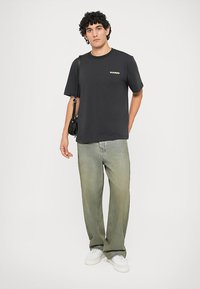 Musta puuvillapaita lyhyillä hihoilla ja etuletolla, yhdistettynä vihreisiin suoralahkeisiin farkkuihin ja valkoisiin lenkkareihin. Malli pitää kädessään pientä mustaa laukkua.