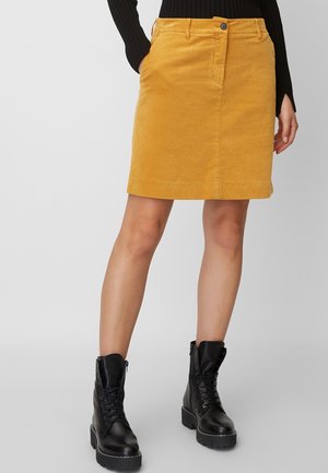 Vrouw die een mosterdgele ribfluwelen rok, een zwarte top met lange mouwen en zwarte veterlaarzen draagt, staand tegen een grijze achtergrond.