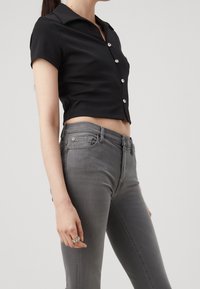 Camisa negra corta con mangas cortas, que presenta un cierre frontal con botones y botones brillantes, combinada con jeans de mezclilla gris ajustados.