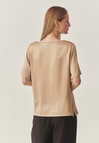 Beige Seidenbluse mit kurzen Ärmeln, Rundhalsausschnitt und Seitenventilationsschlitzen, kombiniert mit dunkler Hose. Glatte Struktur, minimale Nähte.