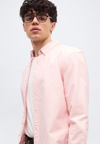 Jeune homme aux cheveux bouclés et foncés portant des lunettes, vêtu d'une chemise rose pâle boutonnée par-dessus un t-shirt blanc, devant un fond uni.