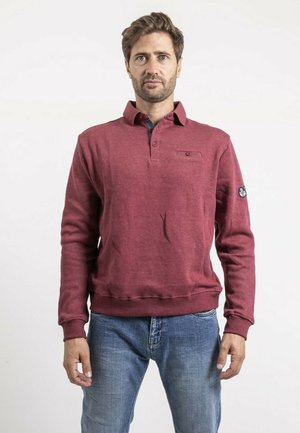 Homme debout face à l'avant, portant un pull-over bordeaux à manches longues avec un col à boutons et un jean bleu, sur un fond blanc.