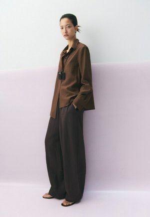 Mujer de pie contra una pared lavanda y blanca, con una camisa marrón de manga larga, pantalones anchos oscuros, chanclas y un collar con colgante negro.