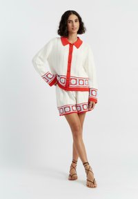 Breisoutfit met een witte cardigan met rode kraag en accenten, versierd met blauwe en rode bloemenpatronen, gecombineerd met bijpassende shorts.