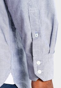 Chemise boutonnée bleu clair avec un tissu texturé, des poignets à un bouton et un ourlet courbé avec des fentes latérales.